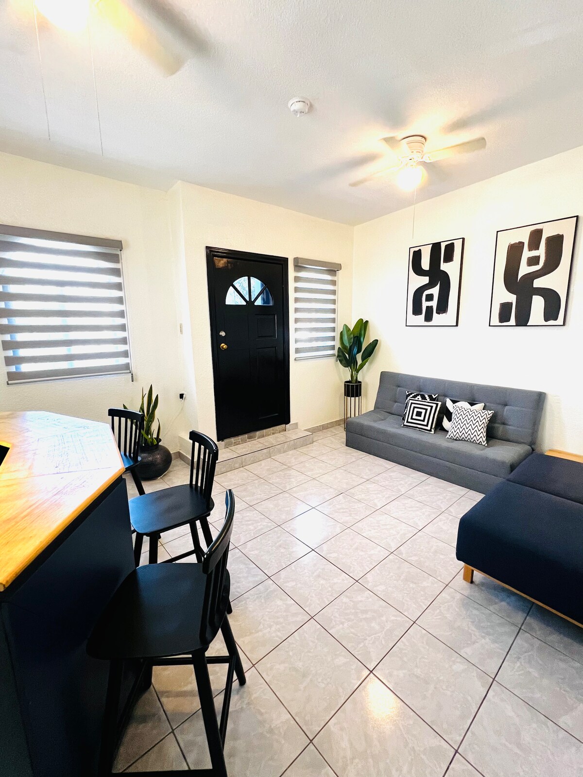 A bright living area is observed, featuring a gray sofa and a wooden breakfast bar with black chairs. A front door with a circular window adds character, while two large framed art pieces decorate the walls. Natural light filters through the window blinds.