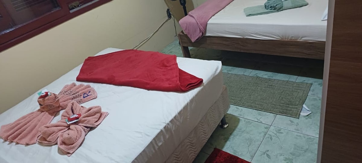 A shared room contains two beds: a spacious bed topped with a red blanket and a smaller bed with a green towel at the foot. Various towels are arranged neatly on the beds. A small rug is positioned on the floor, contributing to the room's functionality.