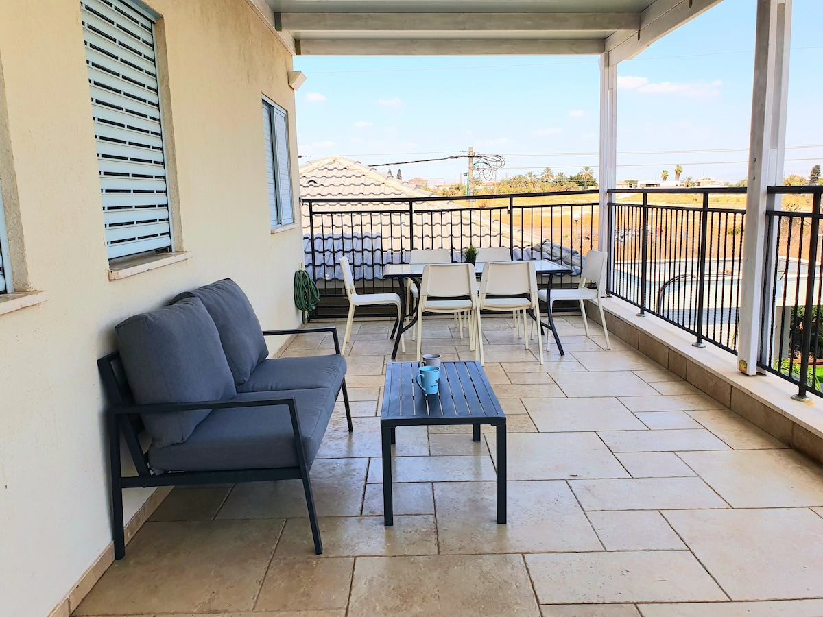 A spacious balcony is furnished with a gray sofa and a black coffee table, complemented by white dining chairs. The area is surrounded by a metal railing, offering views of the serene landscape beyond, with a clear blue sky visible overhead.