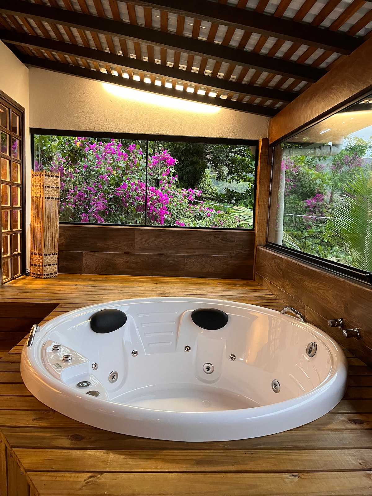 A relaxing space is presented featuring a round whirlpool bath situated on a wooden deck. Large windows display vibrant pink flowers and lush greenery, creating a serene natural backdrop. Soft lighting enhances the tranquil atmosphere of the room.
