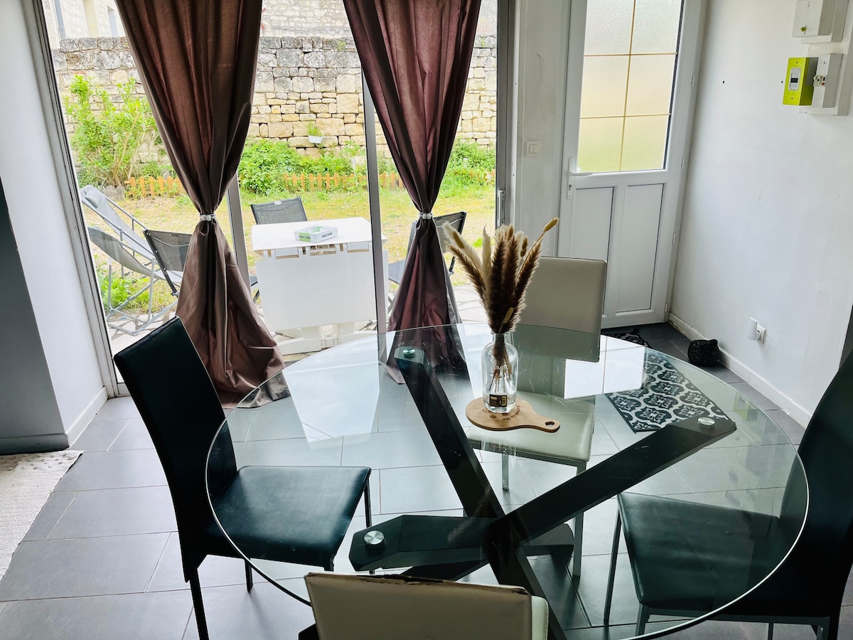 A dining area features a glass table surrounded by black chairs. Natural light filters through large windows, which are adorned with brown curtains. A vase with dried plants is centered on the table, while a view of the garden is visible through the sliding doors.