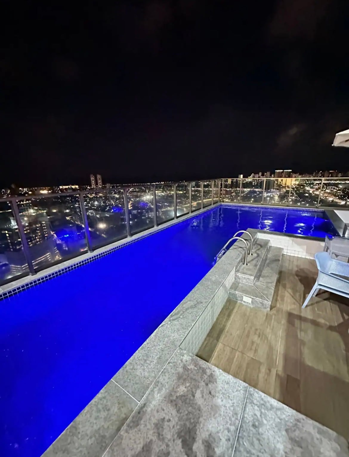 A rooftop pool glows with a deep blue hue, surrounded by a glass barrier that offers unobstructed views of the city skyline. A spacious deck features a seating area with modern chairs, enhancing the tranquil ambiance of the space.
