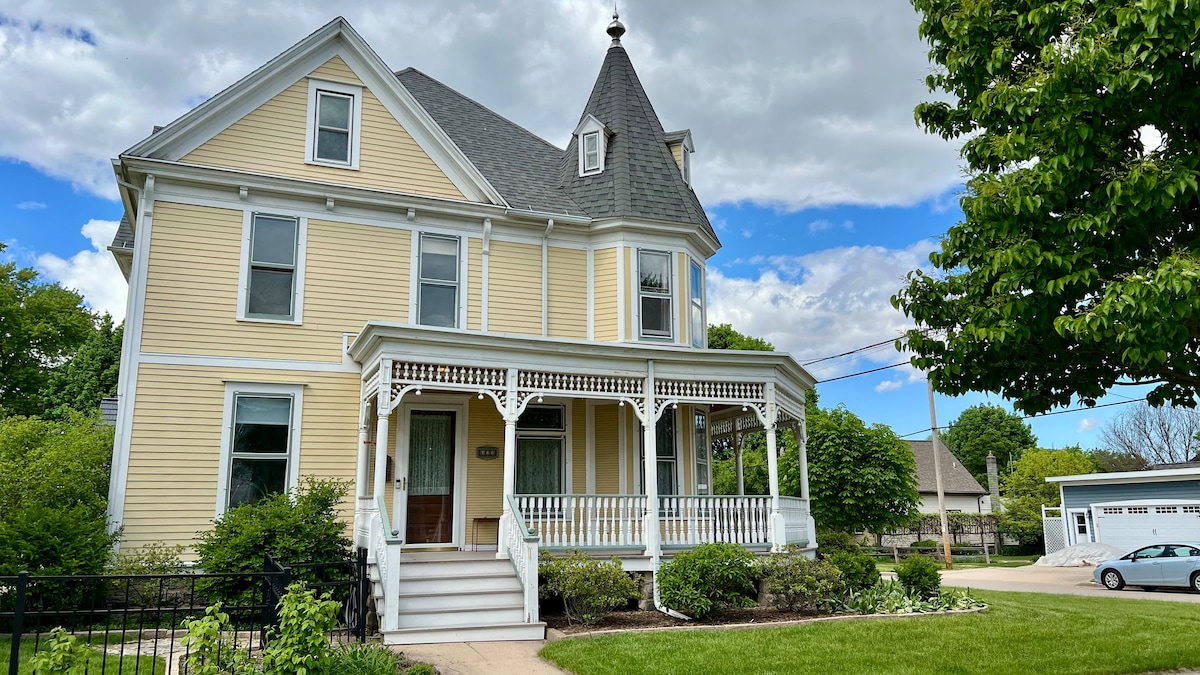 A well-maintained historic Victorian house stands proudly with its distinctive turret and wrap-around porch. The exterior is painted in soft yellow with white trim, complemented by vibrant green landscaping and a clear blue sky above. The inviting entrance features decorative railings and a stone pathway.