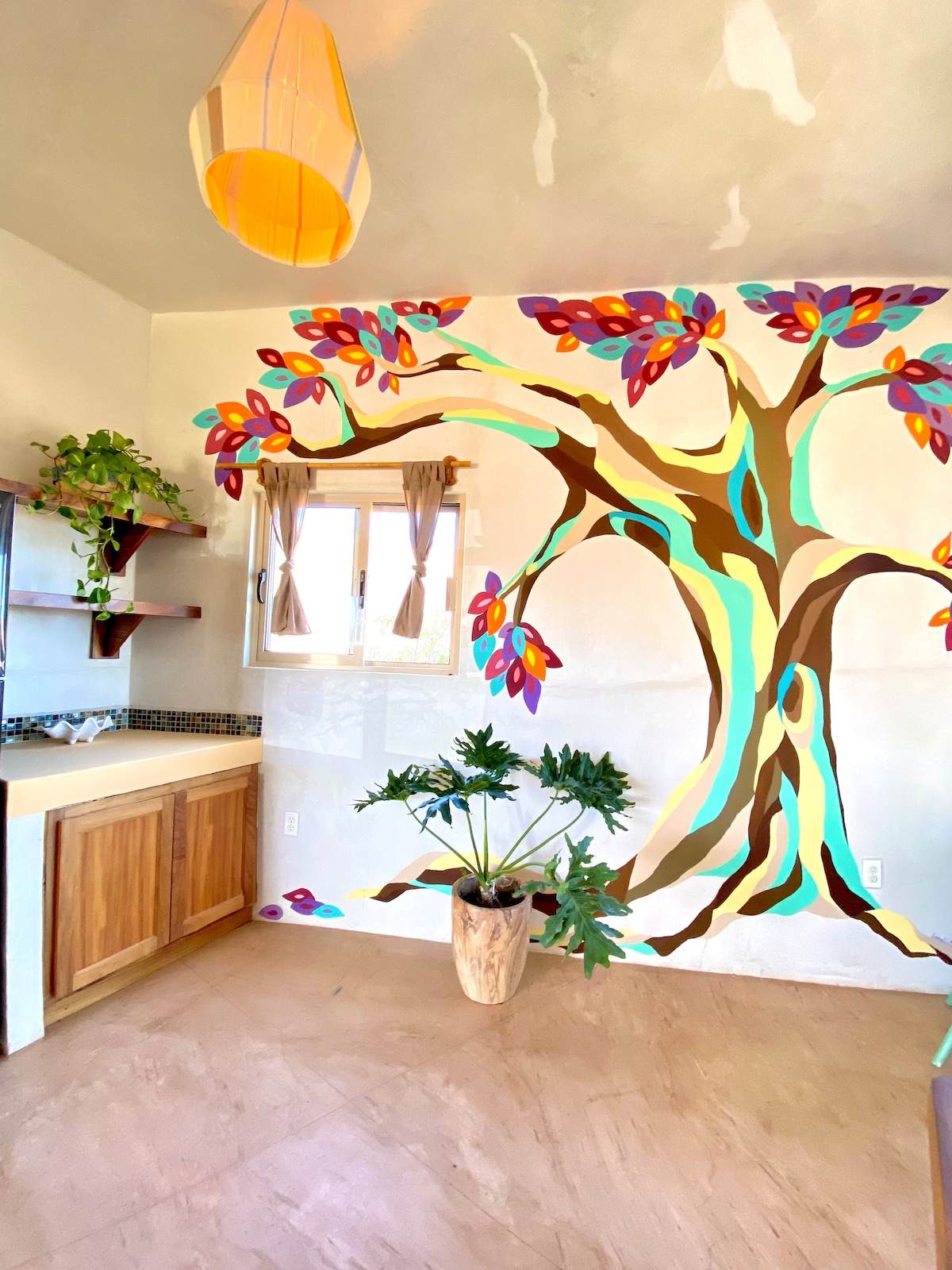 A vibrant mural depicting a colorful tree adorns one wall, complemented by a large potted plant. Natural light enters through a window with light-colored curtains, and a kitchen area features wooden cabinetry and a simple countertop, creating a lively yet serene atmosphere.