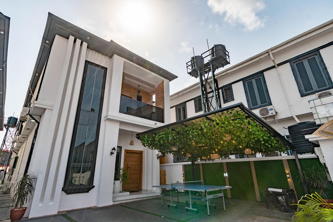 The exterior of the building features modern architecture with large glass windows allowing natural light. An outdoor area is highlighted by a table tennis setup beneath a green trellis, creating a relaxed and inviting environment. Clear blue skies and sunlight enhance the overall ambiance.