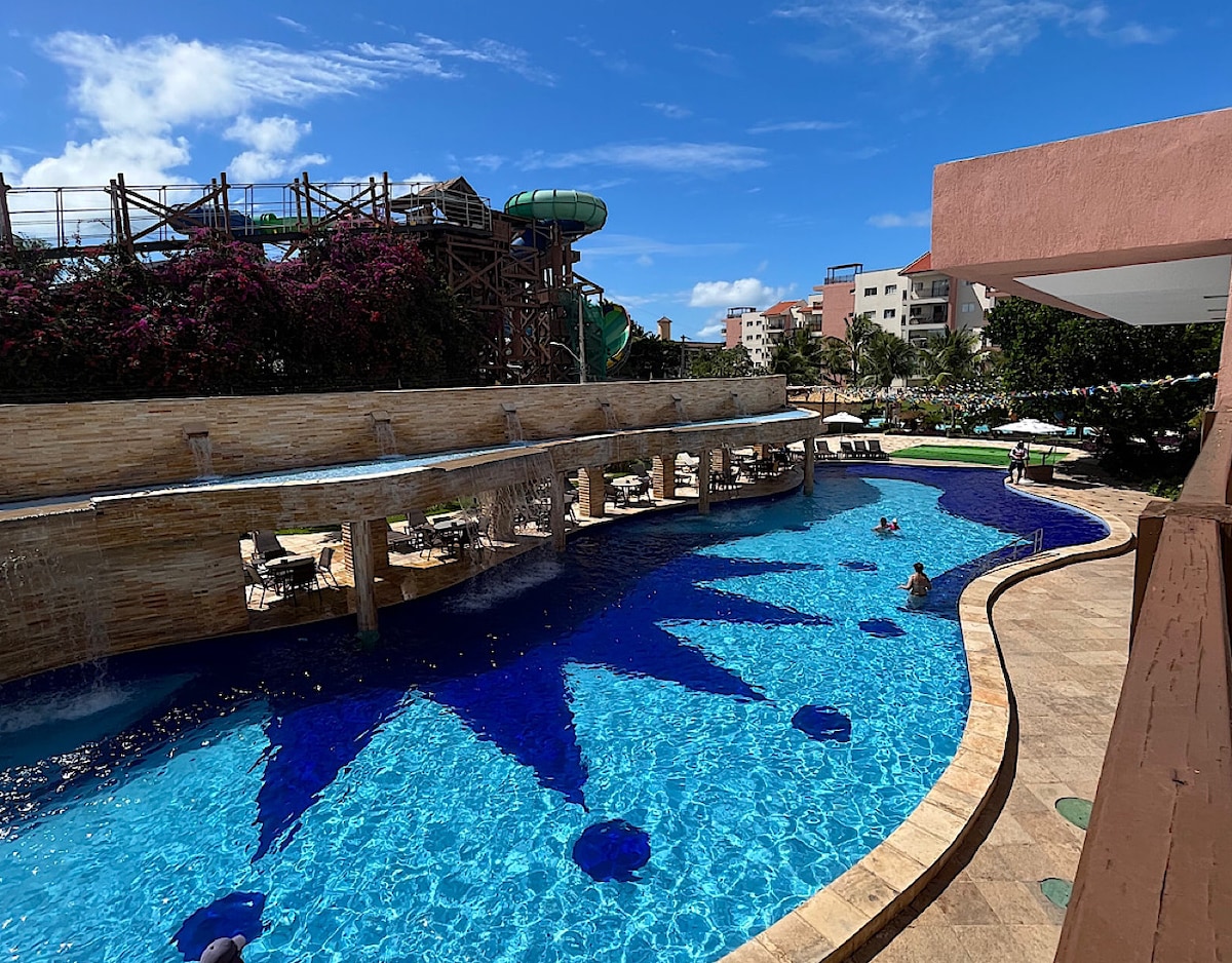 A resort-style pool is featured, showcasing deep blue water and playful geometric patterns on the surface. Lush greenery surrounds the pool area, while lounge chairs and tables are positioned along the water. A water slide is visible in the background.