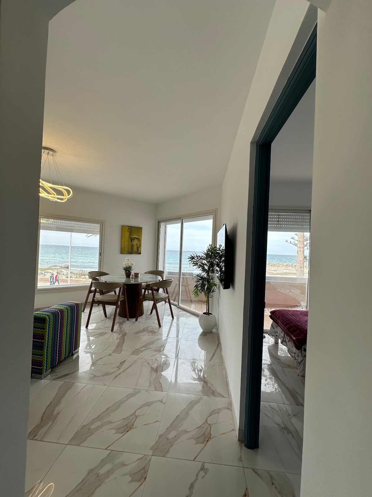 A bright corridor leads to a sunlit living space overlooking the beach. Large windows frame ocean views, while a dining table with chairs sits beside the windows, and a decorative plant adds a touch of greenery.
