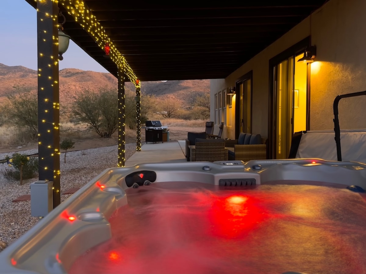 A relaxing hot tub is illuminated with soft red lights, positioned on the back porch. The surrounding area features string lights, enhancing the serene atmosphere. A gas grill and outdoor seating are visible under a shaded area, with desert landscape and mountains in the background.