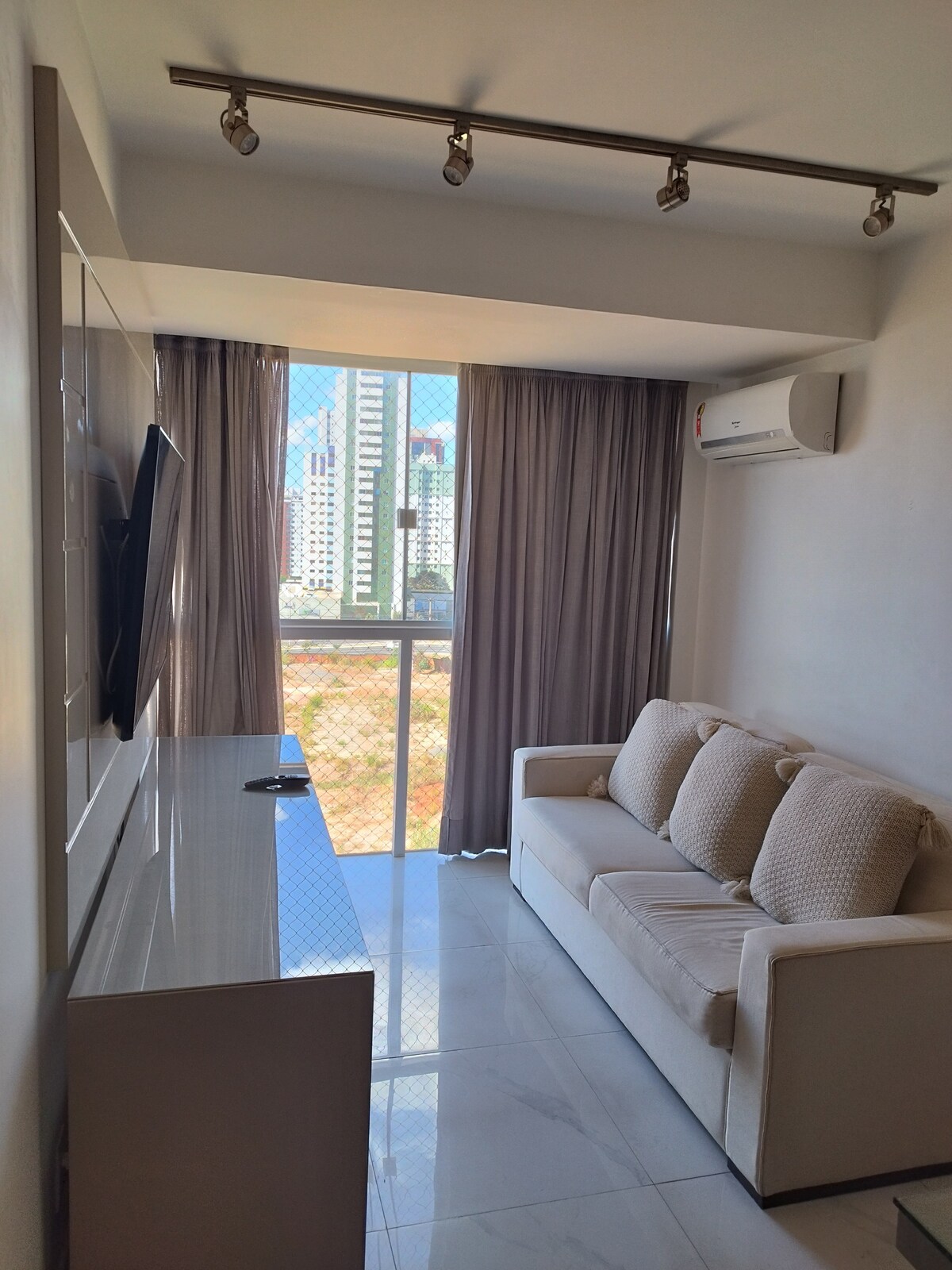 A well-lit living area features a light-colored sofa with textured cushions, positioned in front of a modern television unit. Large glass doors lead to a balcony, offering views of the urban skyline, while gray curtains frame the windows to manage natural light.