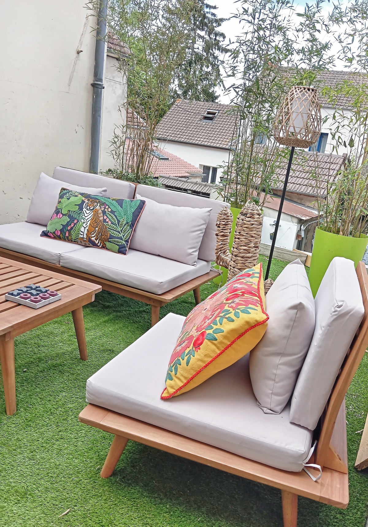 An outdoor seating area is displayed, featuring two comfortable couches adorned with colorful throw pillows. A small wooden coffee table sits between them, creating a cozy space. Lush greenery and decorative lanterns add to the serene atmosphere of the terrace.
