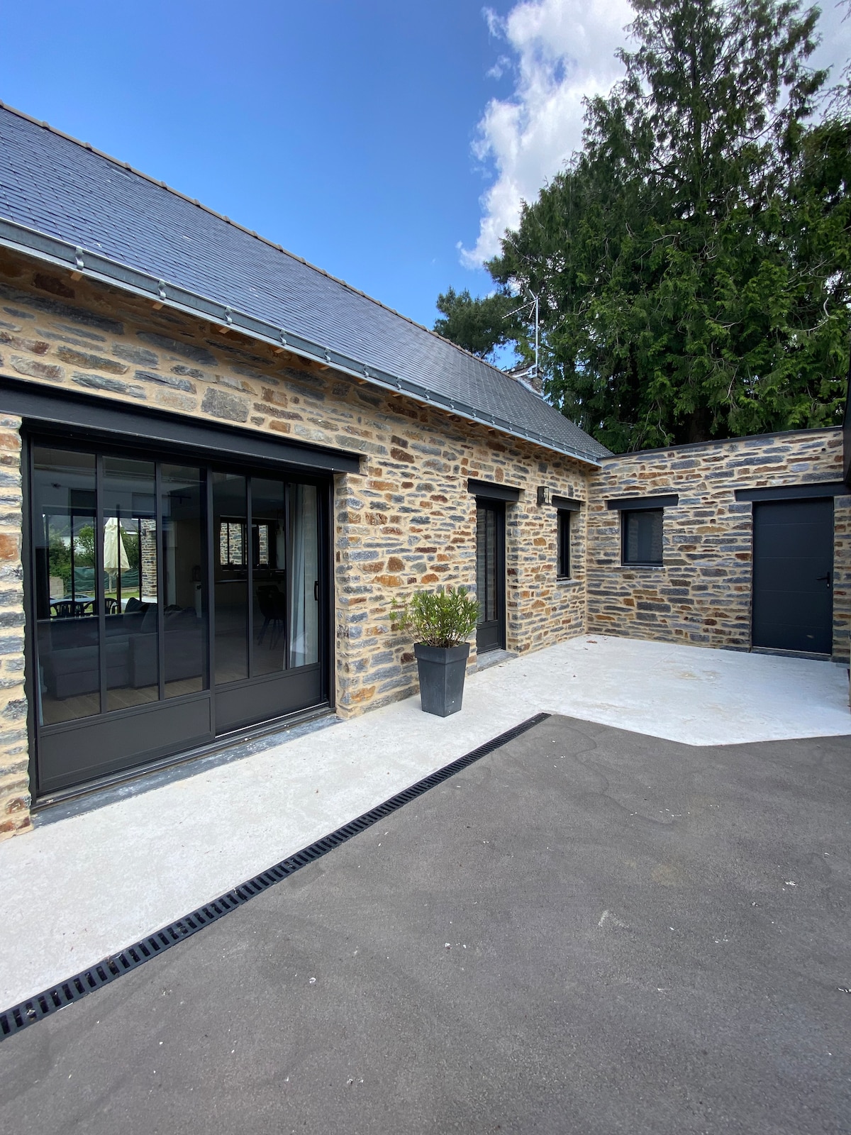 The exterior of a recently renovated stone house is depicted, featuring a smooth concrete area. Large glass doors provide views into the interior, framed by dark window frames. Potted plants add a touch of greenery next to the entrance, while a pathway leads to the adjoining structure.