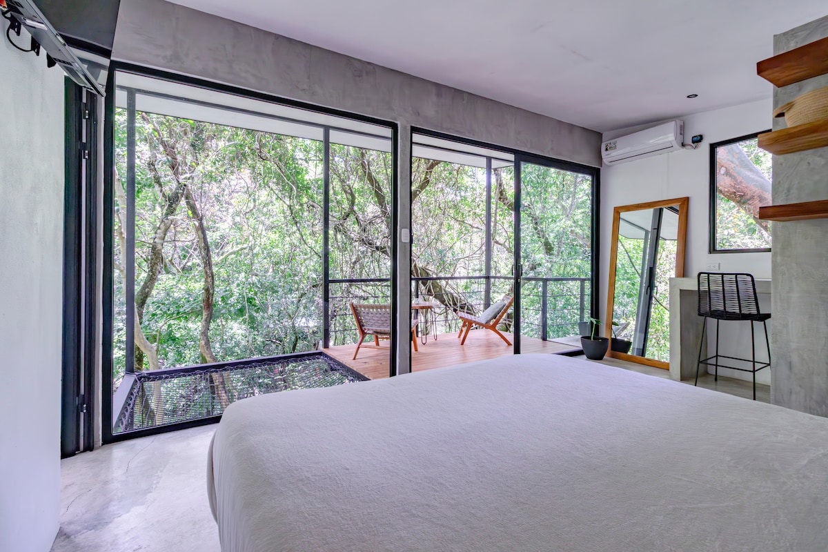 A spacious studio is presented, featuring large glass walls that provide a view into the surrounding greenery. The area includes a comfortable queen bed, a small seating arrangement on a balcony, and a natural-inspired decor that integrates with the outdoor environment.