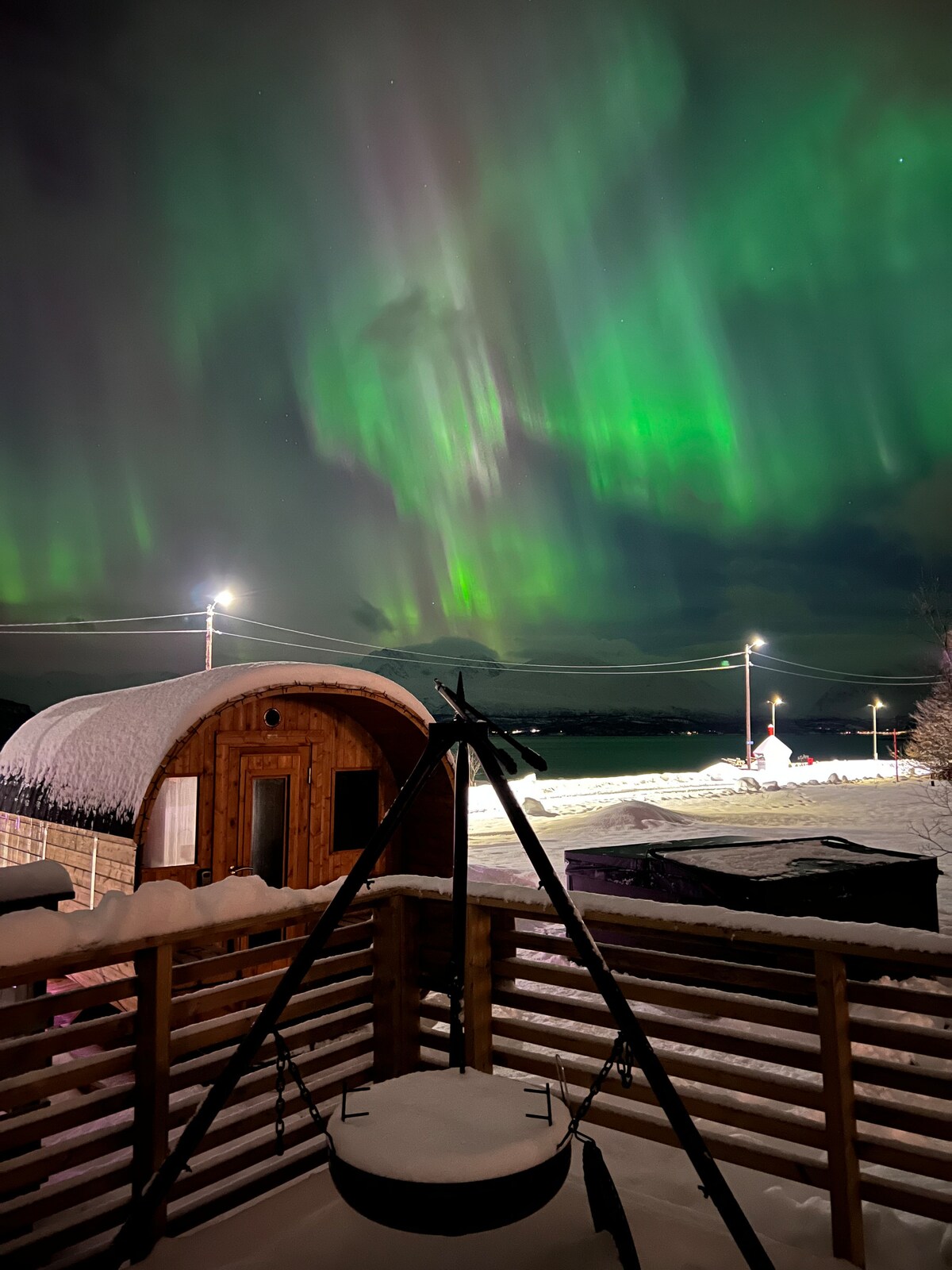 The image depicts the enchanting northern lights illuminating the sky with vibrant greens. In the foreground, a wooden structure, likely a sauna or storage unit, blends harmoniously with the snow-covered landscape. A nearby tripod fire pit is surrounded by fresh snow, enhancing the serene atmosphere.