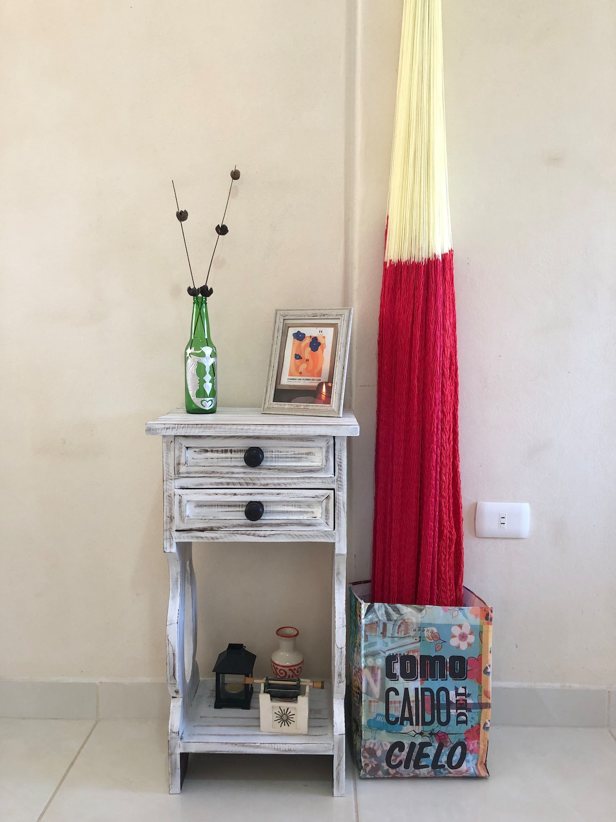 A small white side table with a drawer is positioned against a light wall. It displays a colorful framed artwork, a green glass bottle with decorative twigs, and a lantern. Next to it, a red and yellow woven hanging decoration leans alongside a decorative bag featuring vibrant lettering.