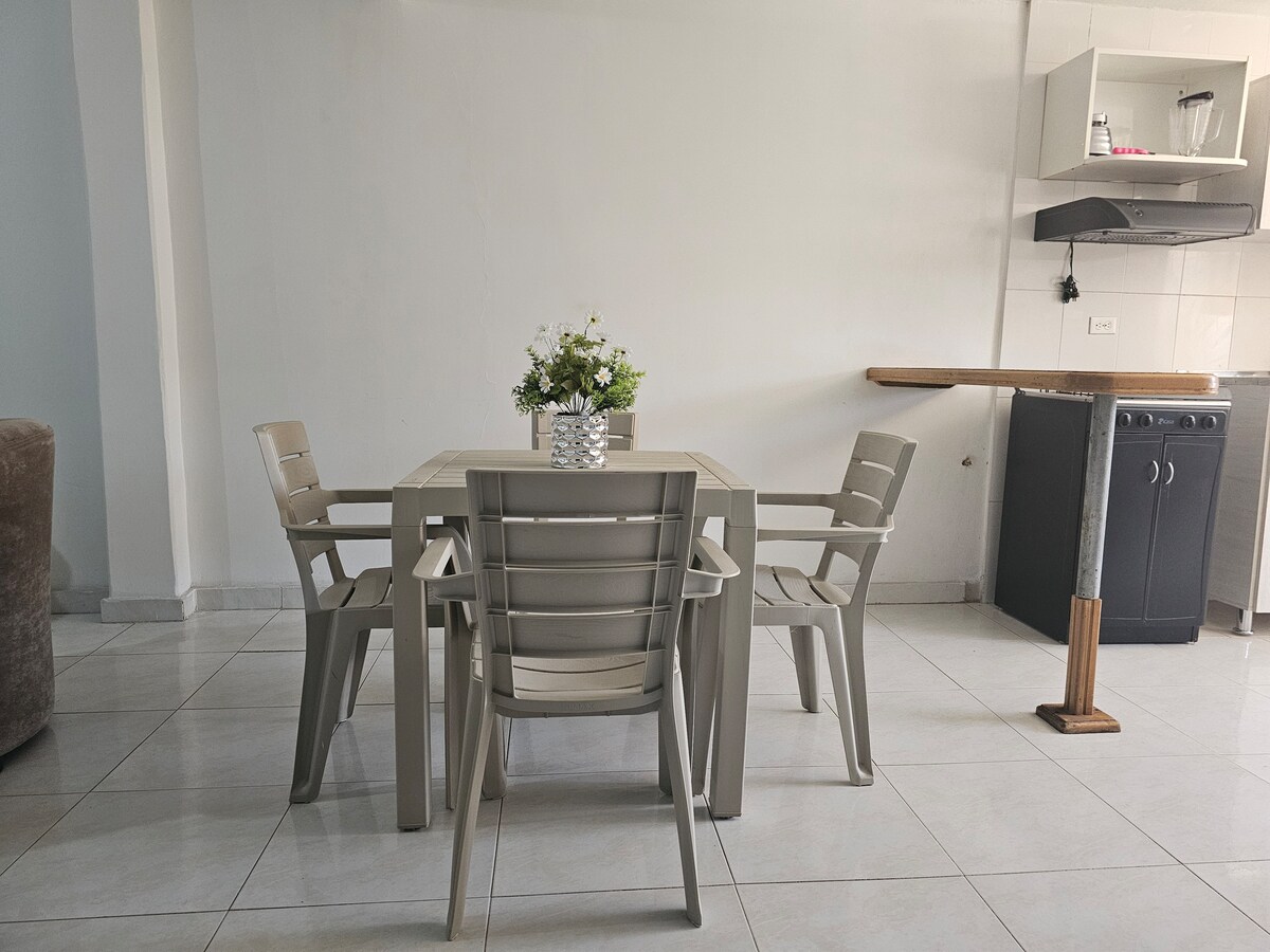 A dining area is shown, featuring a square table surrounded by four light-colored chairs. A small vase with flowers rests on the table. The space includes a minimalist kitchen area in the background, with a small sink and stove set against a white wall.