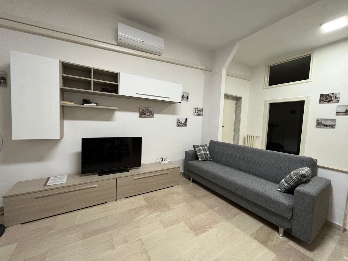 A modern living area is displayed with light-colored tiled flooring. A comfortable gray sofa is positioned against the wall, accompanied by decorative pillows. A flat-screen TV is mounted on a minimalistic entertainment unit, while wall-mounted shelves and framed images add subtle decor.