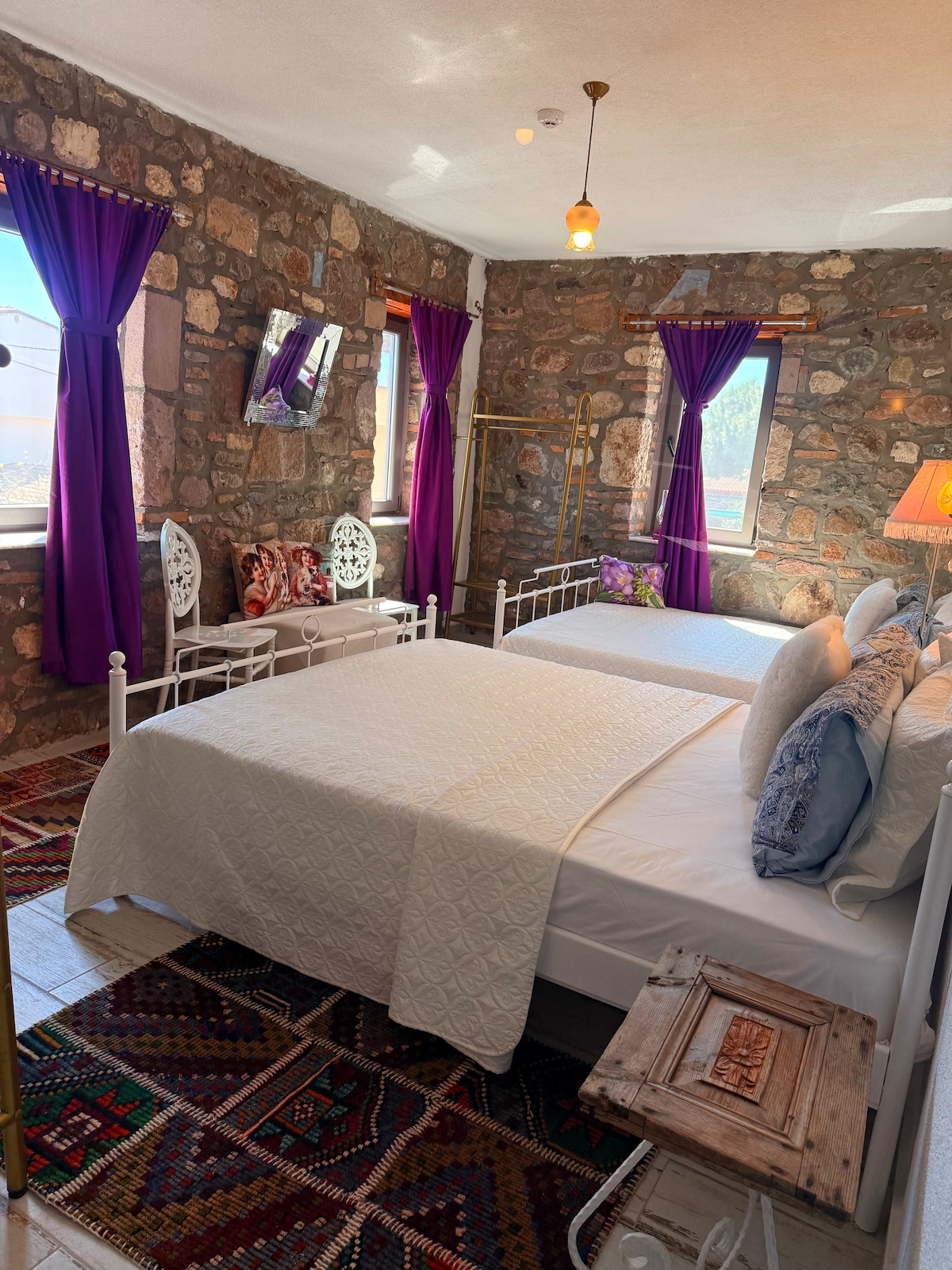 A cozy bedroom features two inviting beds, adorned with crisp white linens and patterned throw pillows. Stone walls and vibrant purple curtains complement the decor. A wooden side table and a colorful rug enhance the space, providing a warm and welcoming ambiance.