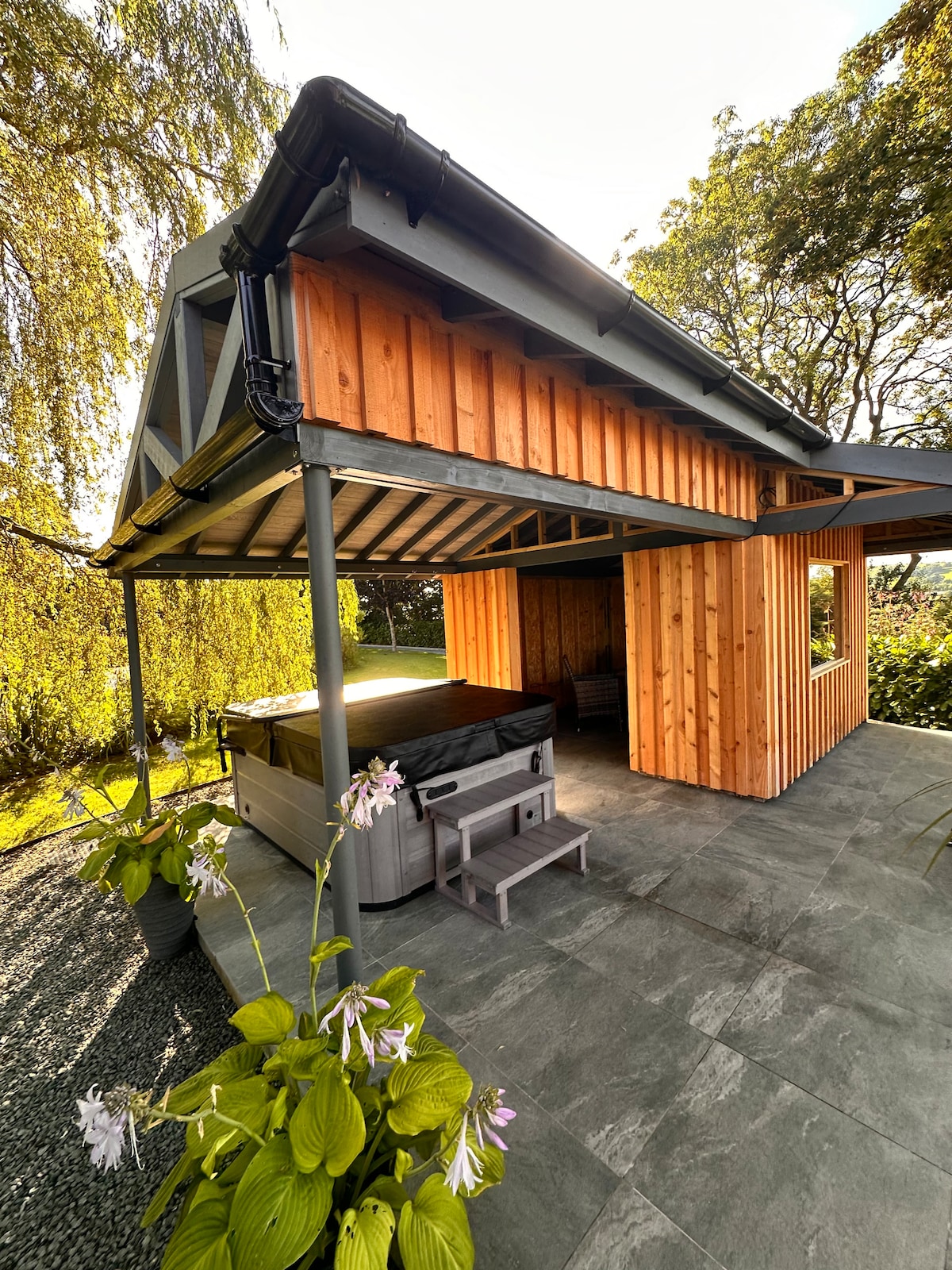 An outdoor area is showcased featuring a covered hot tub sheltered by a modern roof. Adjacent to the tub, a small seating arrangement is visible. Lush greenery and flowers frame the scene, enhancing the natural setting around the hut.