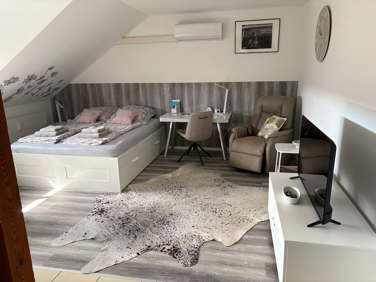 A modern room features a spacious bed with neutral bedding set against a textured wall. A comfortable chair and desk are arranged near a window, while a soft area rug lies across the floor. A TV stand with a television completes the inviting space.