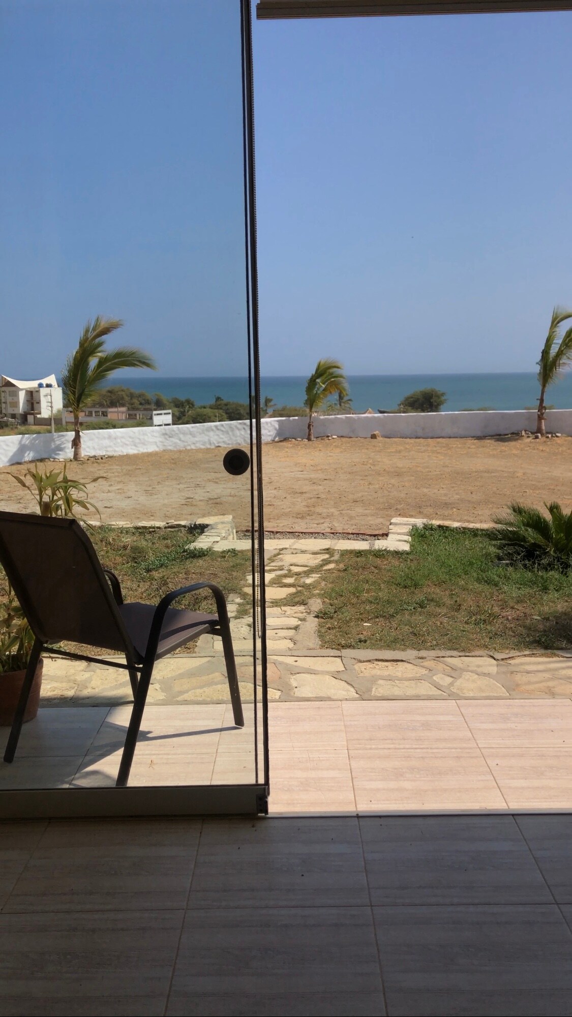 A clear view is presented through sliding glass doors, showcasing a sandy area leading to the ocean. A single chair is positioned on a tiled floor, with palms swaying gently in the foreground and a calm sea visible in the background under a blue sky.