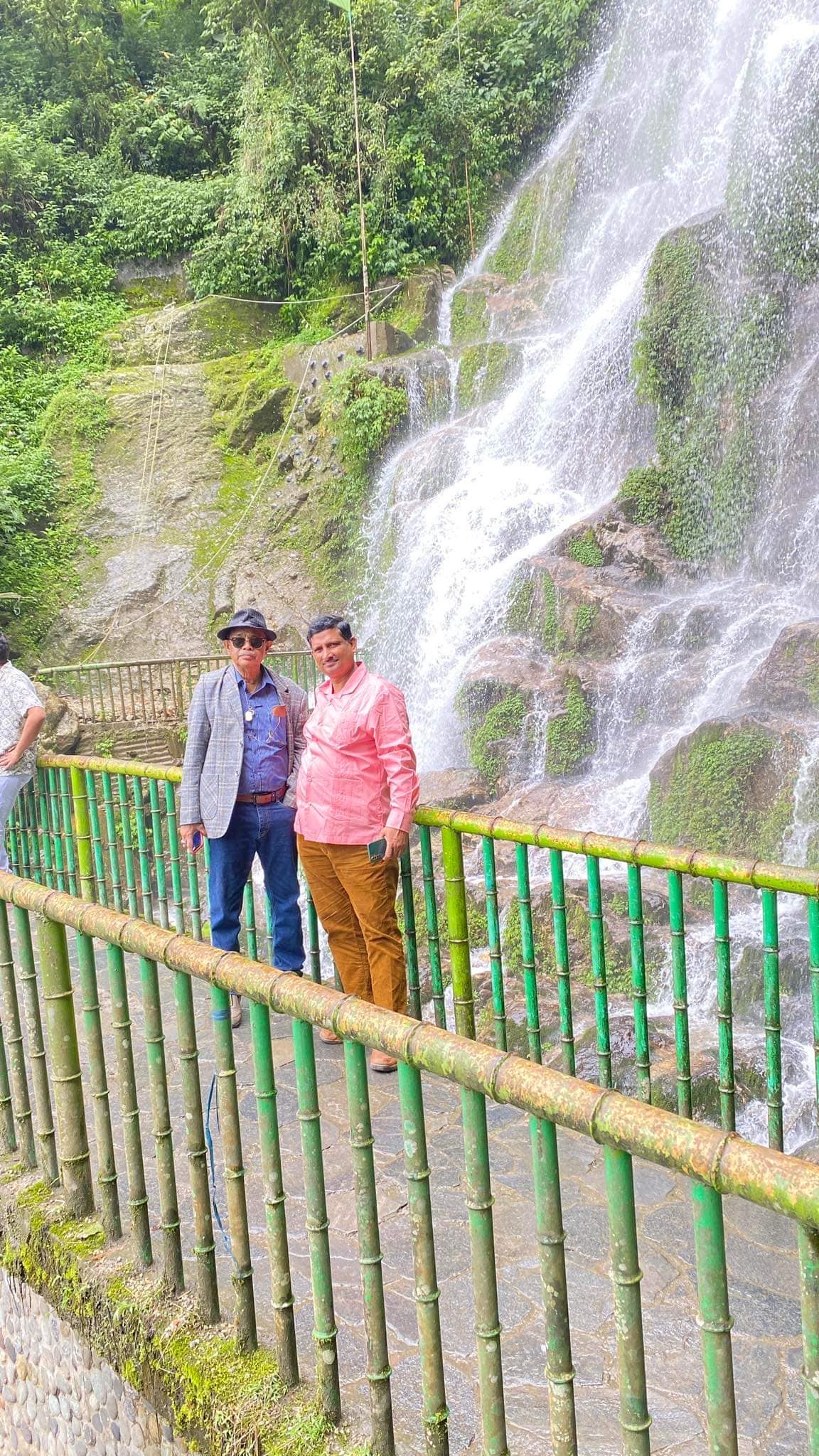 A vibrant waterfall cascades down rocky cliffs, surrounded by lush greenery. Two individuals stand by a green bamboo railing, smiling and enjoying the natural scenery. The gentle mist from the water glistens in the light, creating a refreshing atmosphere.