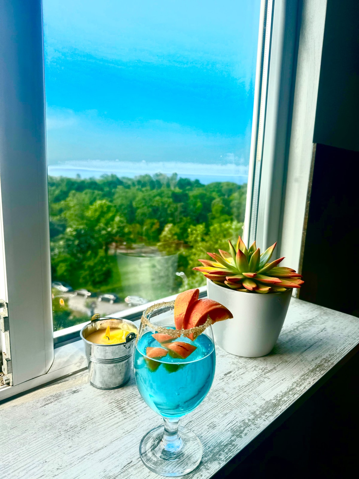 A drink served in a glass with a bright blue hue is positioned on a windowsill alongside a small potted succulent and a candle in a metal holder. The window frames a view of green trees and a glimpse of the sea beyond.