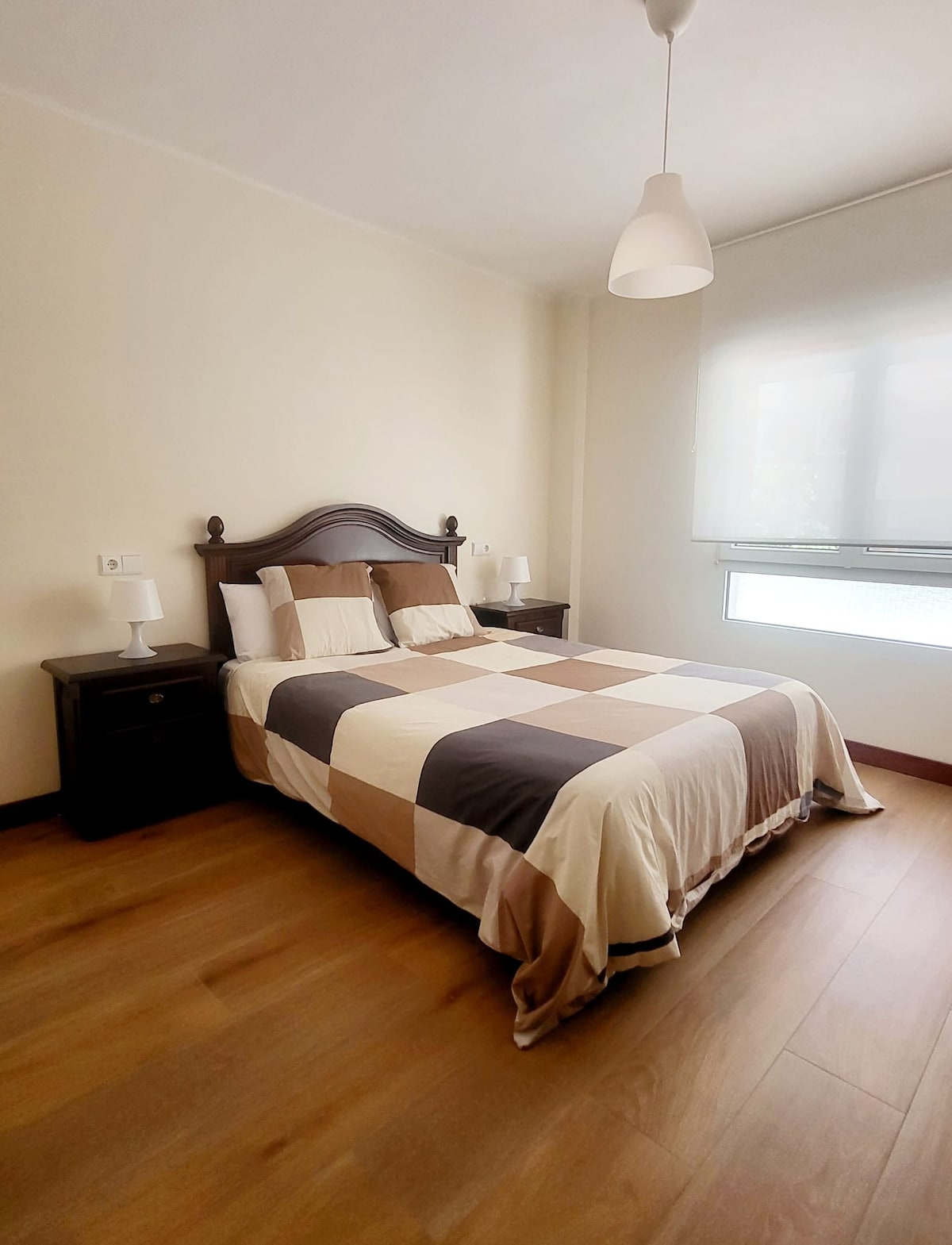 A spacious bedroom features a large bed with a quilted cover that includes a patchwork design. Two nightstands with lamps flank the bed, and a large window allows natural light to fill the room. Light wood flooring complements the neutral wall color.