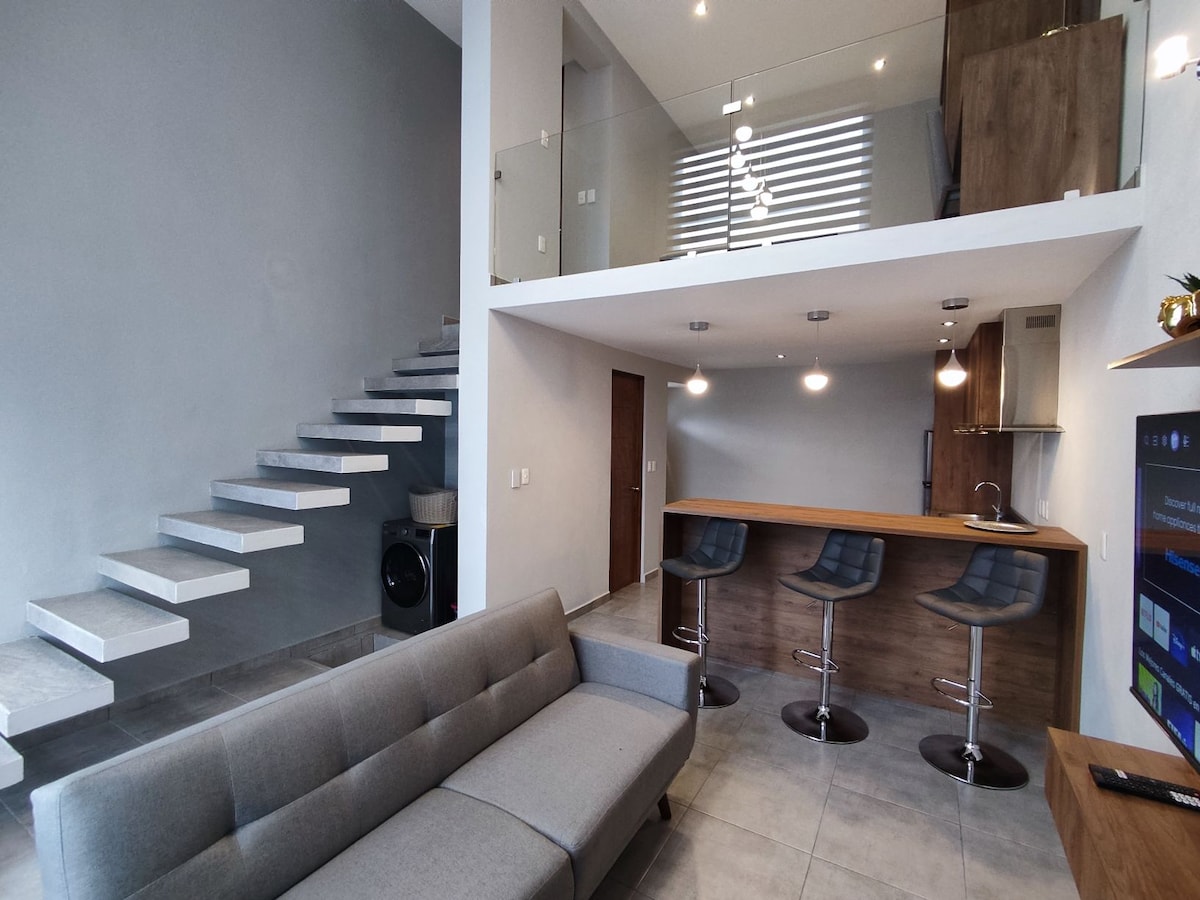 The open-concept living area showcases a modern gray sofa and a sleek kitchen bar with three high stools. A staircase with minimalist design leads to the lofted bedroom above, while natural light brightens the space through large windows.