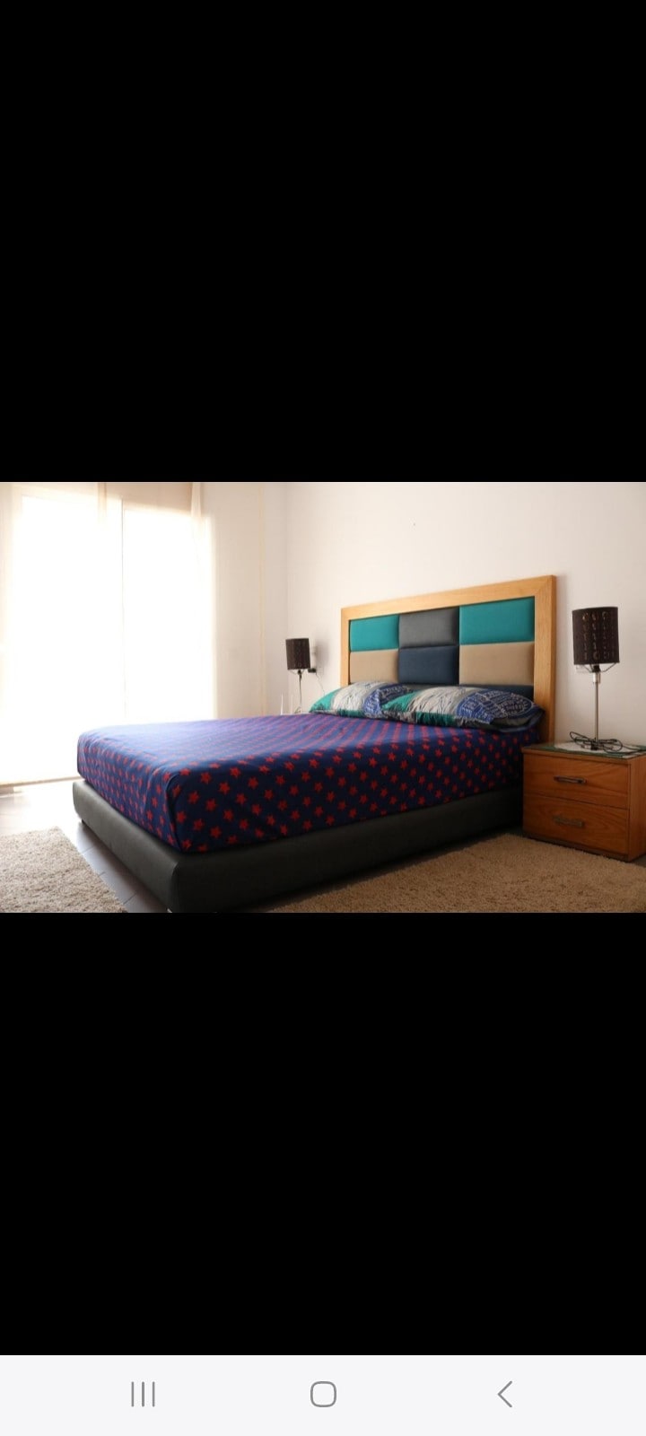A bedroom is presented with a spacious bed dressed in a blue and red polka dot comforter. Natural light flows in from large windows, illuminating the space. Two bedside lamps on either side complement a wooden nightstand.