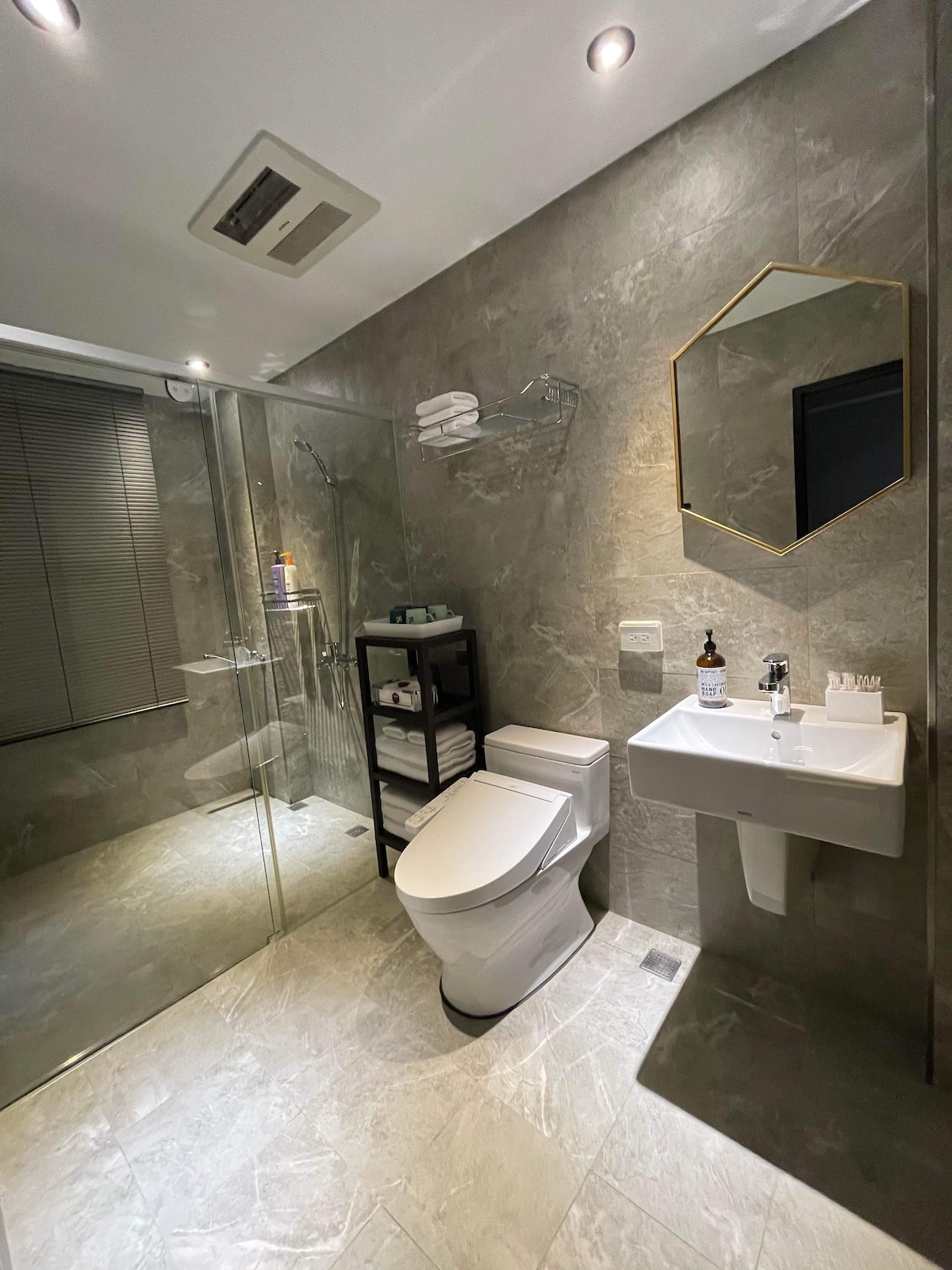 A modern bathroom is displayed, featuring a walk-in shower with clear glass doors. A sleek toilet is positioned next to a compact sink, above which an octagonal mirror is mounted. Towels are neatly arranged on a black shelving unit against the wall.
