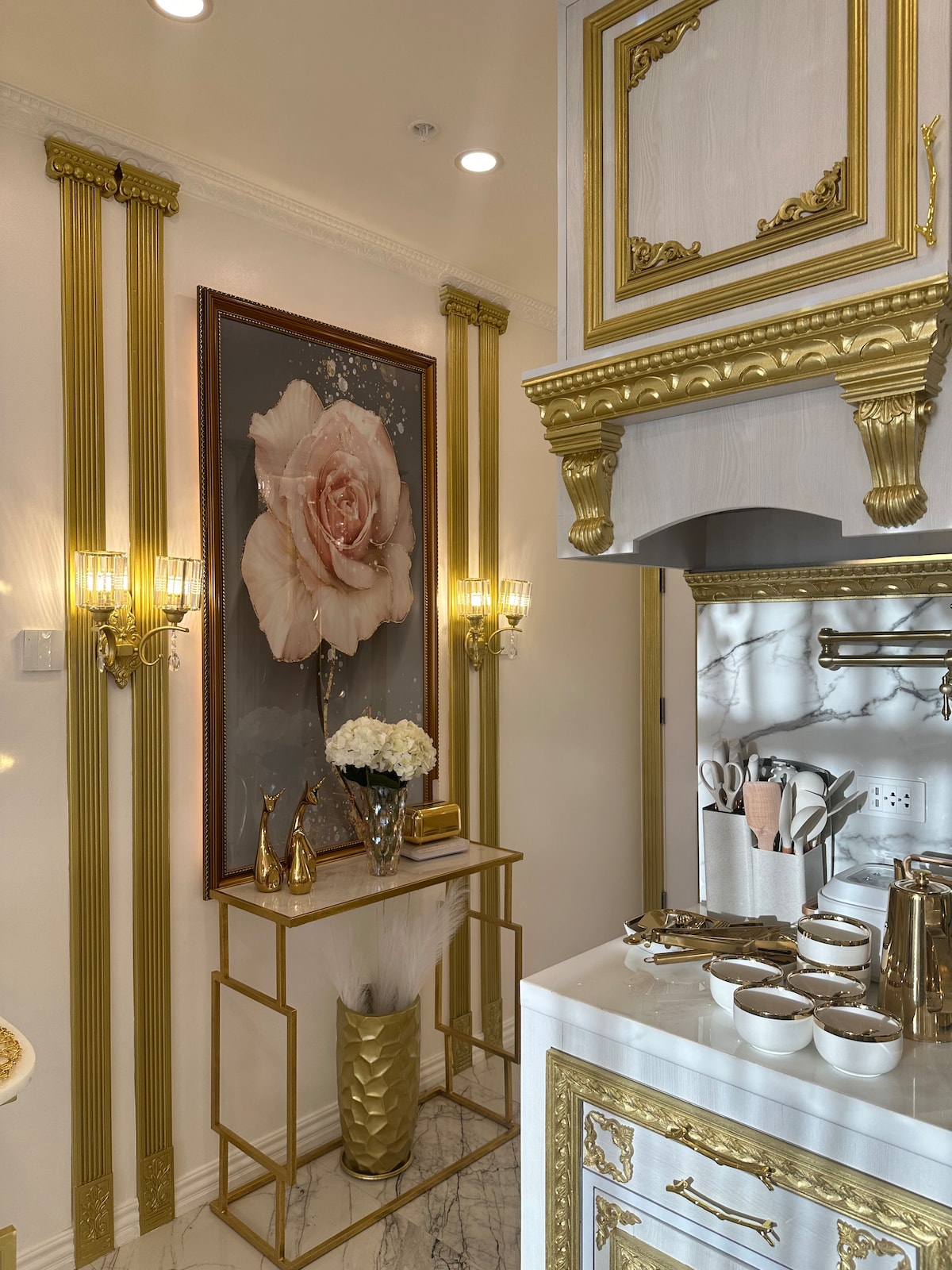 A chic kitchen space is showcased with ornate gold detailing on cabinetry and walls. A large floral artwork adds a touch of elegance. Decorative lighting fixtures illuminate a sleek countertop, which is complemented by stylish kitchenware and soft textures in vases.