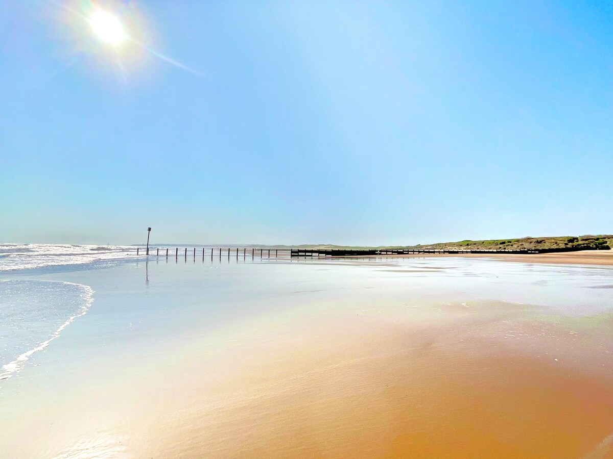 A sandy beach stretches toward the horizon under a clear blue sky, with gentle waves lapping at the shore. In the distance, a wooden pier extends into the ocean, complemented by tufts of grass on the nearby dunes.