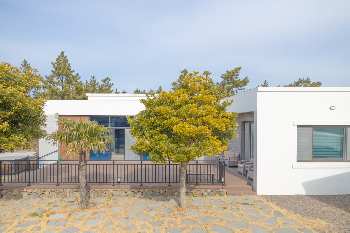 The exterior view showcases a modern home framed by lush greenery, featuring a spacious wooden deck. A pathway made of large stones leads to the entrance, while clear glass doors enhance the connection to the outdoors.