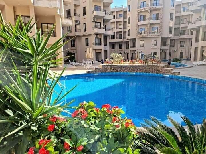 A vibrant pool area is surrounded by lush greenery and colorful flowers. Lounge chairs are positioned near the water, inviting relaxation under the sun. The architecture of the surrounding buildings provides a backdrop of balconies and windows, contributing to a peaceful courtyard atmosphere.