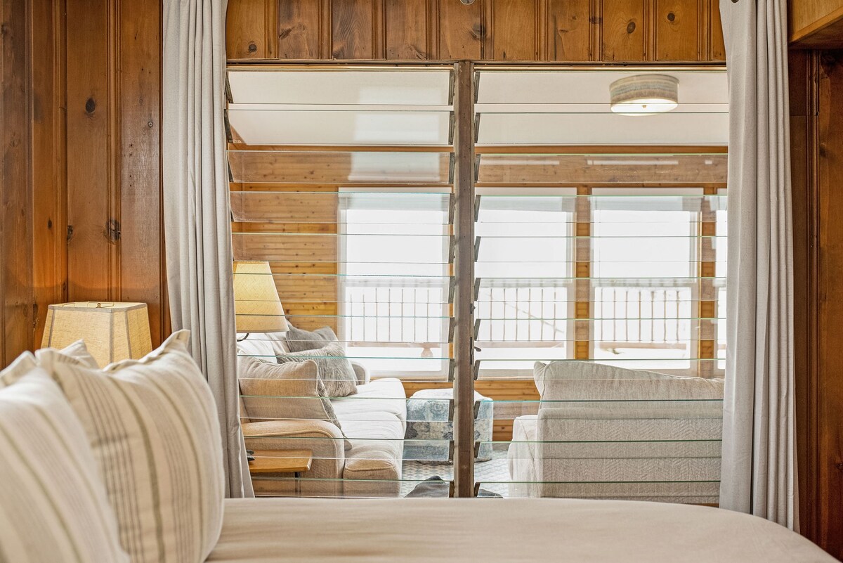 A view through wooden slatted partitions showcases a cozy family room featuring soft couches and lamps. Natural light floods the space, highlighting the warm wood paneling and comfortable atmosphere of the waterfront retreat.