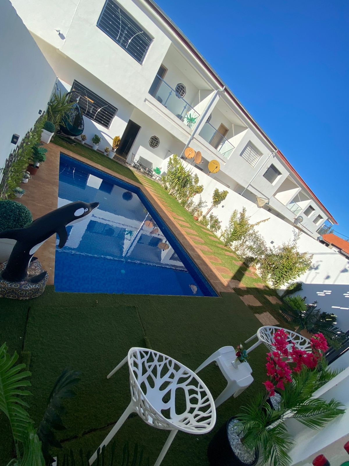 An outdoor area features a private pool surrounded by a grassy landscape. White chairs and a small table sit next to vibrant flowers, complementing the greenery. Two stories of the chalet overlook the pool, with bright blue skies adding to the serene ambiance.