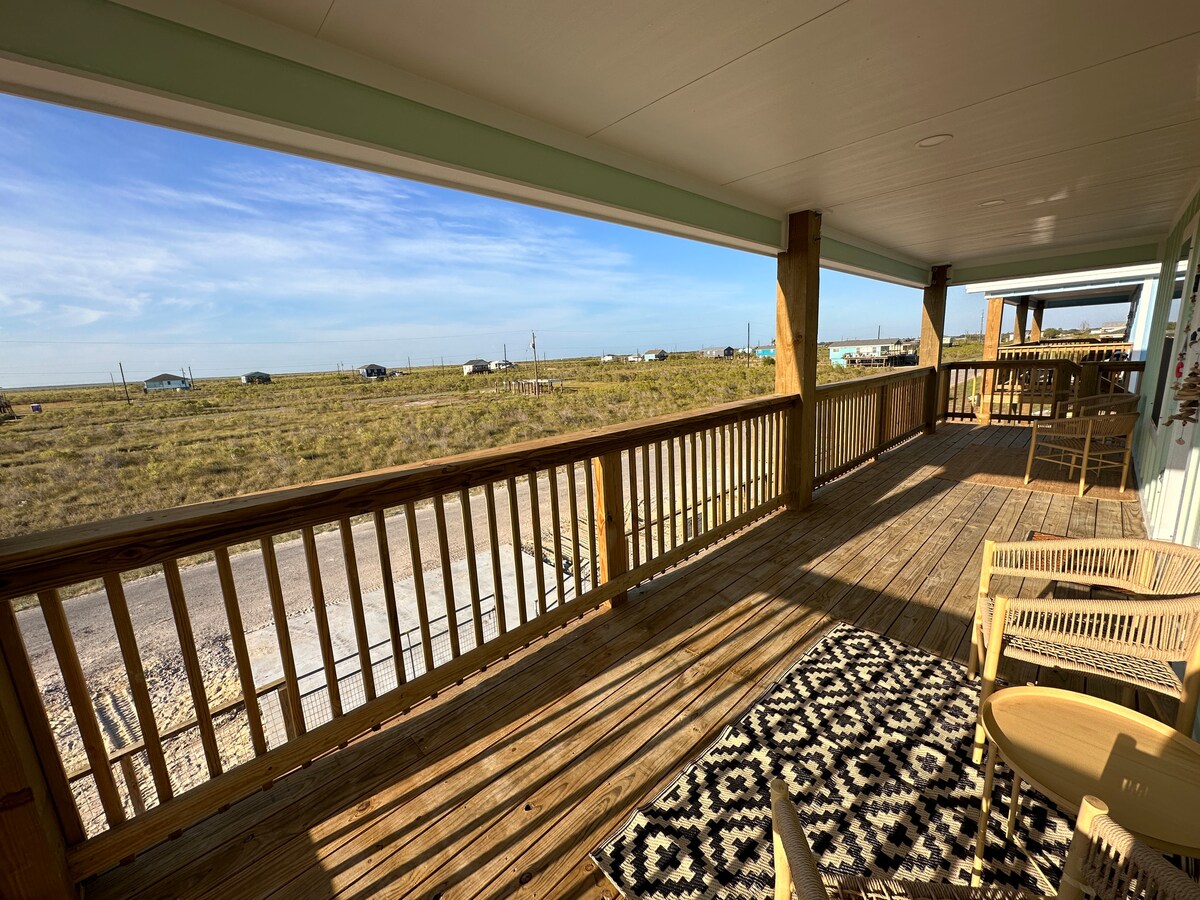 A wooden deck extends from the home, featuring a patterned rug, a small table, and two chairs. Open views of the surrounding landscape and distant vehicles can be seen, with a clear blue sky overhead.