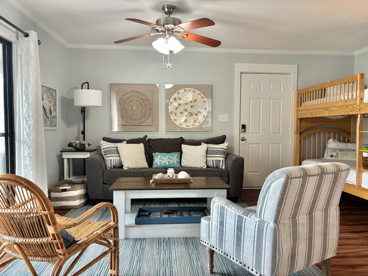 A cozy living area is presented with comfortable seating, including a gray sofa adorned with decorative pillows. A stylish coffee table sits at the center. To one side, a wooden bunk bed is visible, while a chair with a striped pattern adds texture to the space.