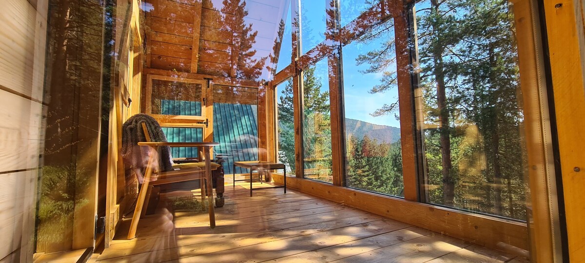 A spacious veranda is surrounded by large glass windows, offering views of the lush forest and mountains. Wooden flooring complements the natural ambiance. A couple of chairs and a small table are arranged for relaxation, inviting guests to enjoy the serene environment.