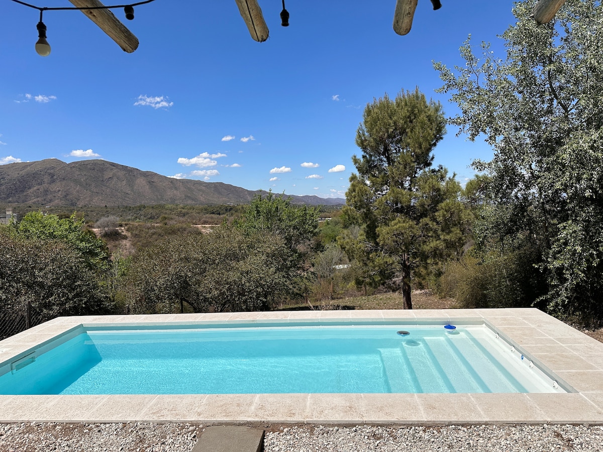 An outdoor pool is depicted, featuring clear water reflecting the sky. Surrounding natural landscapes include rolling hills and green trees under a bright blue sky with scattered clouds. The pool area is framed by a stone deck, enhancing the serene outdoor space.