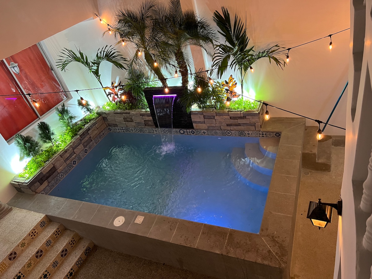 A private pool area is surrounded by tropical plants and illuminated by soft lights. A water feature flows gently into the pool, creating a serene atmosphere. The pool is designed with multiple levels and is encased in decorative stonework.