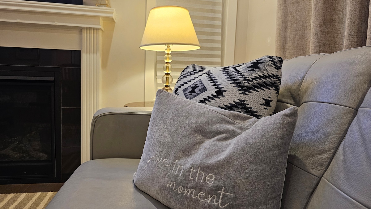 A gray sofa is seen with decorative pillows featuring a patterned design. A warm lamp stands nearby, providing soft lighting. The scene includes a fireplace surrounded by dark tiles, enhancing the cozy atmosphere of the living space.