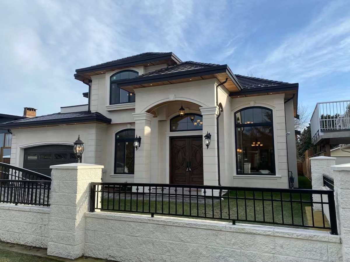 The exterior of a modern two-story house is showcased, featuring a light-colored façade with arched windows. Decorative lanterns flank the grand entrance, which is set beneath an ornate overhang. A low stone wall encloses the front yard, and lush green grass is visible.