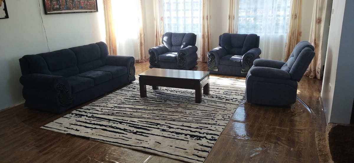 A spacious living area is furnished with a comfortable blue sofa set, including two armchairs and a sectional couch. A dark wooden coffee table sits at the center, surrounded by a striped area rug. Natural light filters through sheer curtains that frame the window.