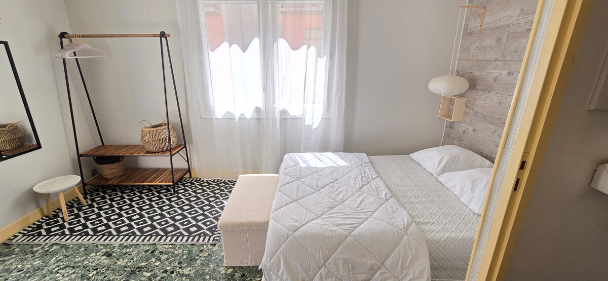 A serene bedroom features a spacious bed with white linens, set against a textured wall. Natural light filters through sheer curtains over two windows, illuminating a patterned rug and a small seating area. A minimalist clothing rack and a shelf are positioned for convenience.