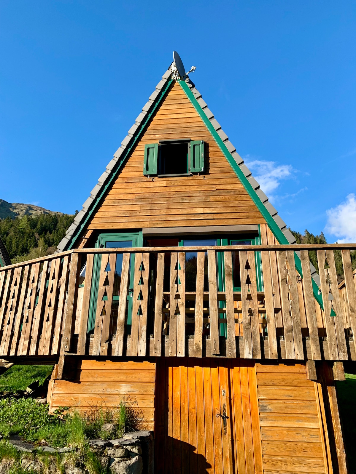 The chalet features a triangular design with wooden siding and green shutters. A spacious wooden balcony extends from the upper level, providing an elevated perspective. Below, the main entrance showcases a natural wood door framed by stone, with a background of clear blue sky and surrounding greenery.