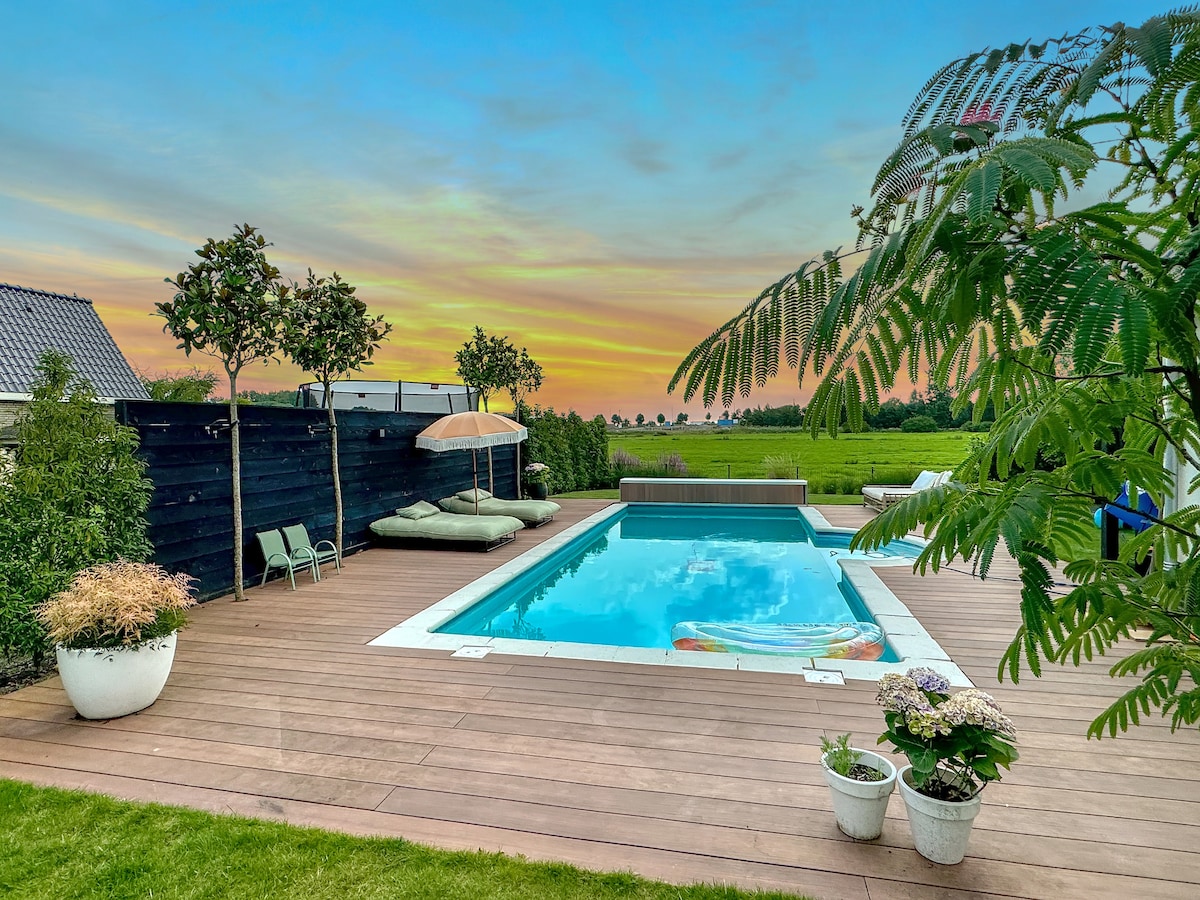 An outdoor space features a heated swimming pool surrounded by wooden decking and green lawn. Comfortable lounge chairs and an umbrella provide shaded seating. Potted plants and small trees add greenery, with a serene sunset sky enhancing the scenic view.