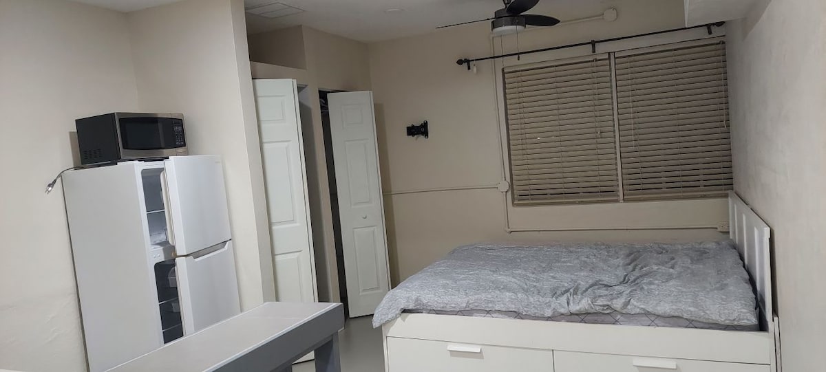 A studio space features a bed with a gray patterned quilt and a ceiling fan overhead. A small dining table is set against a light-colored wall. A kitchenette is equipped with a refrigerator and microwave, with storage cabinets nearby.