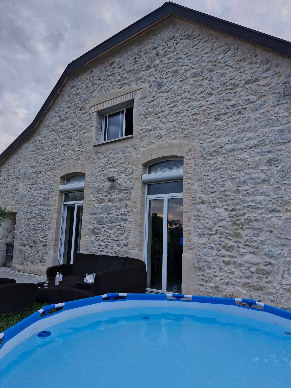 The exterior of a stone house is visible, featuring large windows that invite natural light. A cozy seating area is situated in front of the house, and a small above-ground pool is positioned nearby, enhancing the outdoor living space.