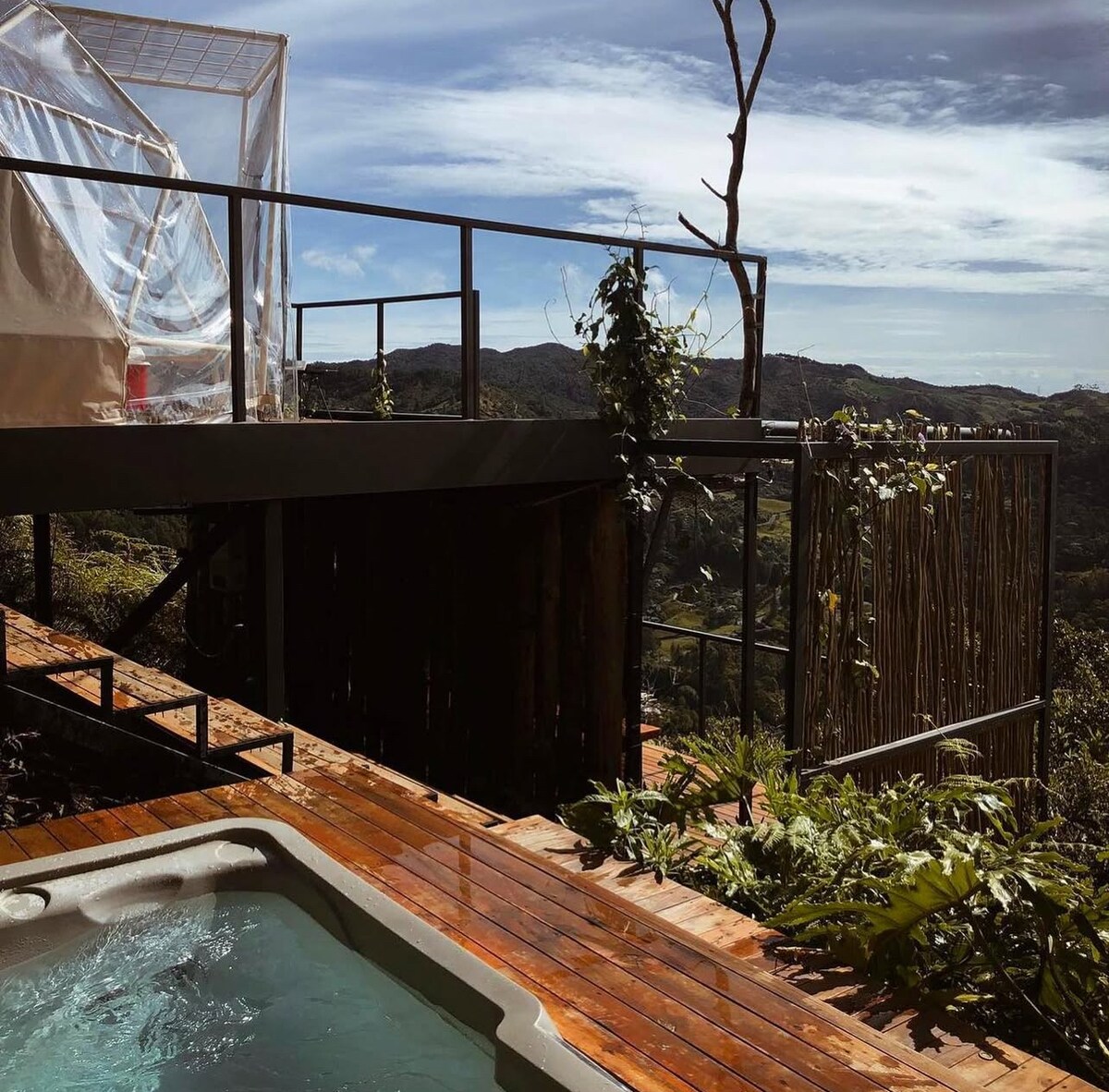 A wooden deck surrounds a hot tub, offering a view of lush greenery and distant hills. Nearby, a glamping structure features a transparent canopy, integrating nature with the living space. The sky is clear, reflecting natural light on the serene setting.